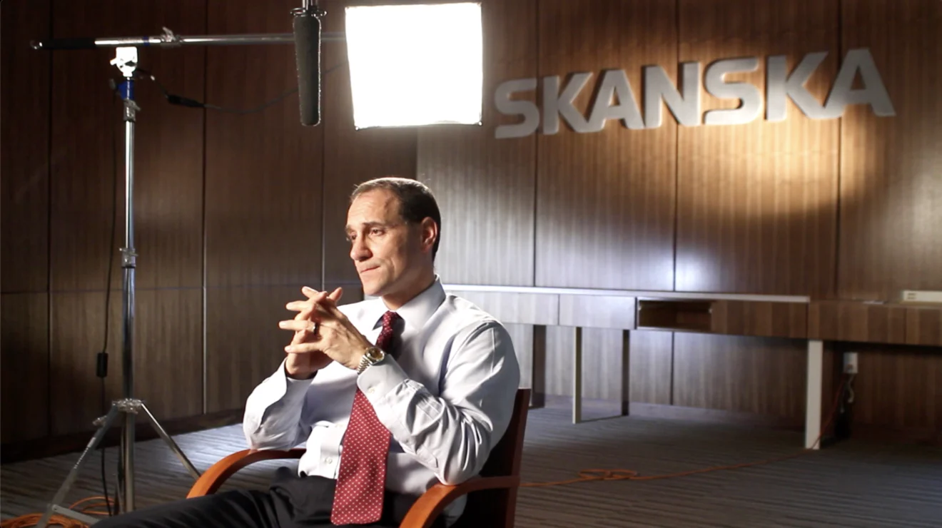 Executive being filmed for an on-camera interview in front of a lit Skanska logo wall, with boom microphone and lighting rig visible